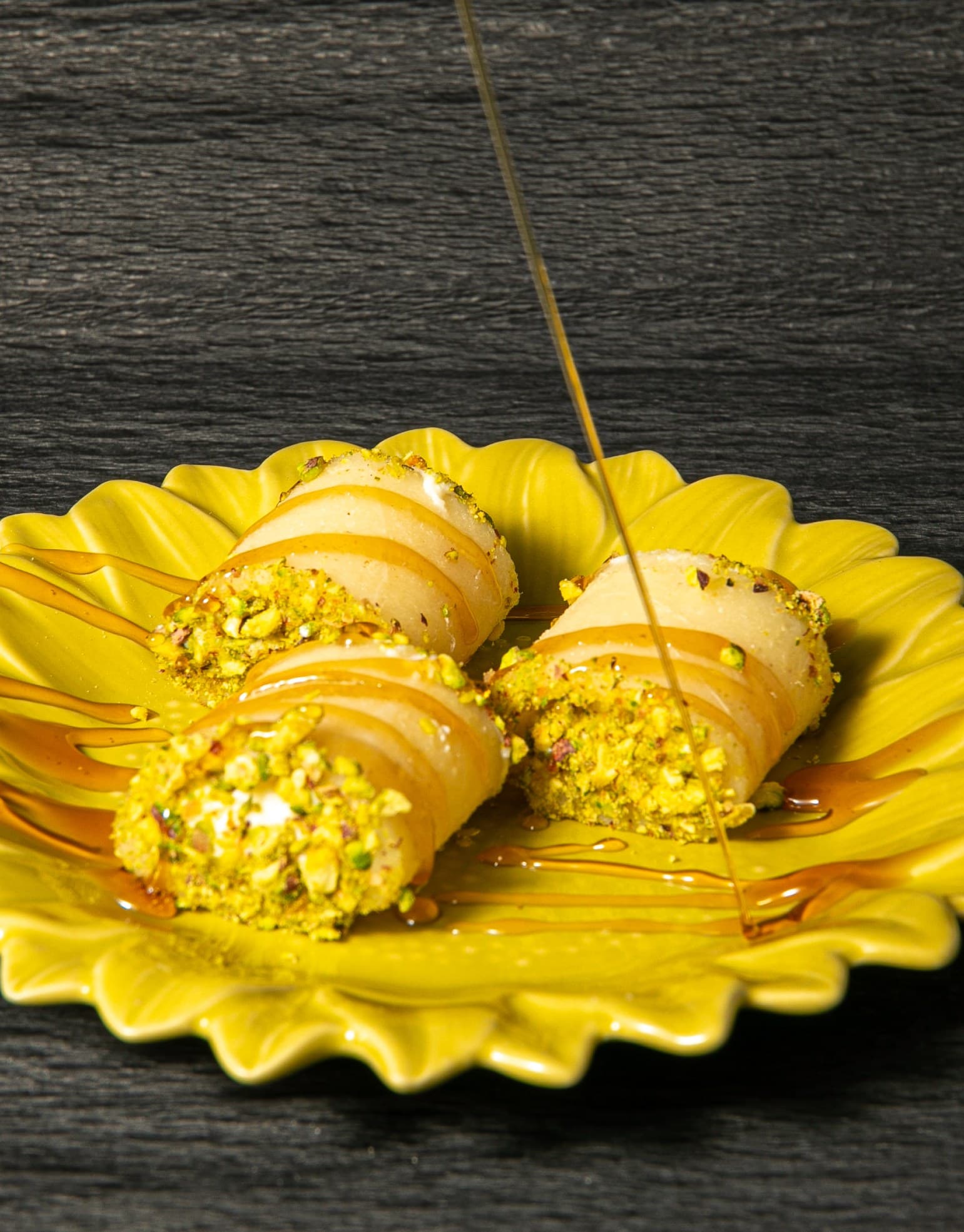 Pistachio-crusted cream rolls on a bright yellow flower-shaped plate, drizzled with honey syrup over dark wood.