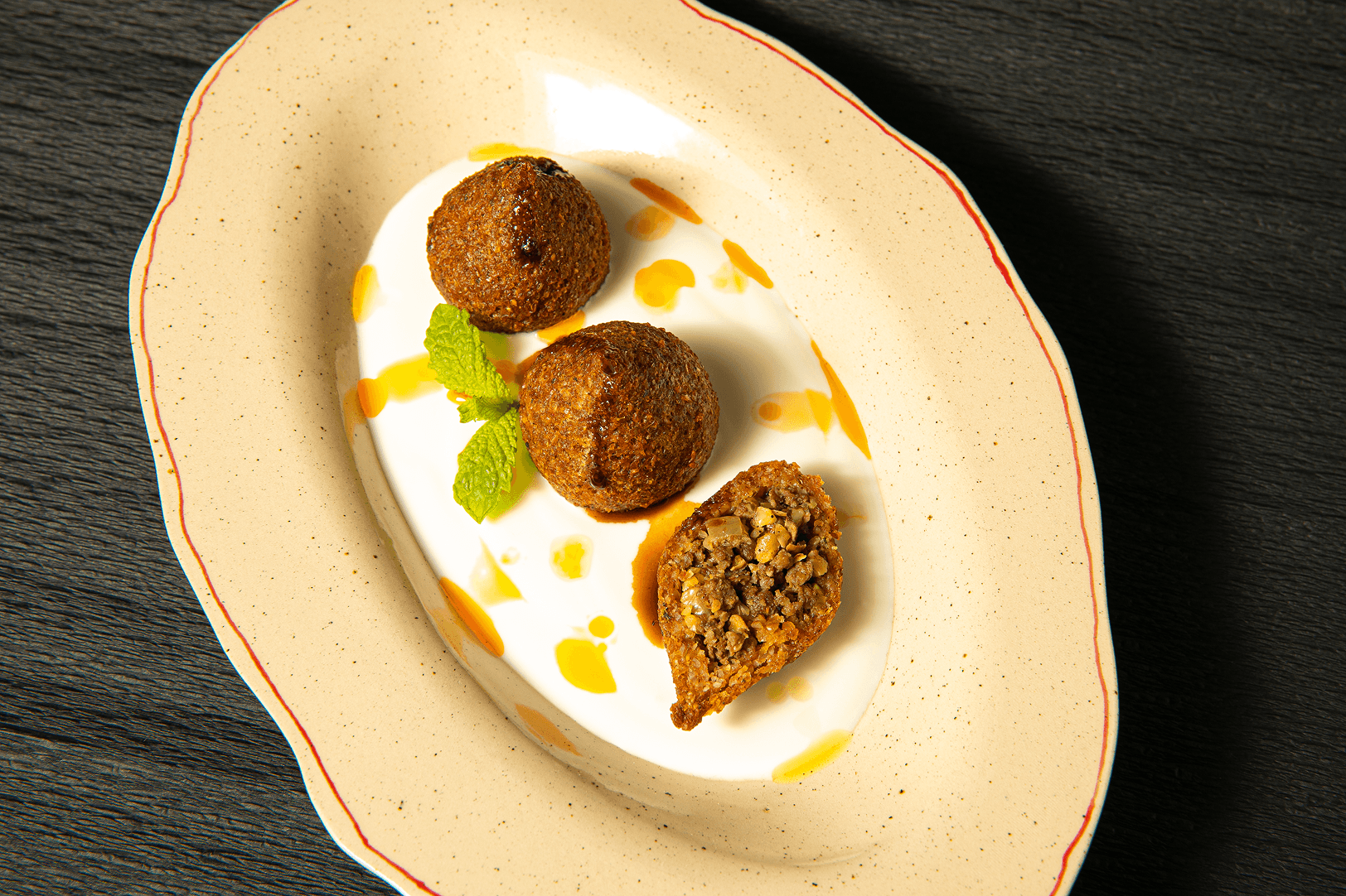 Fried kibbeh resting on a swoosh of yogurt, one halved to reveal its spiced filling, garnished with fresh mint.