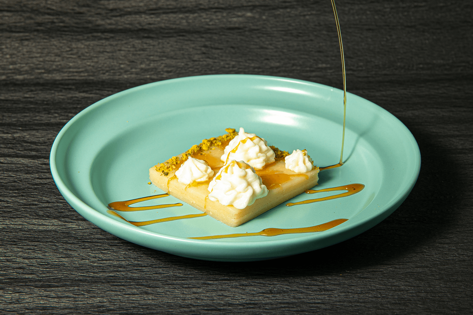 A square of semolina cake topped with cream quenelles and crushed pistachio, finished with a pour of honey syrup, on a mint-green plate.