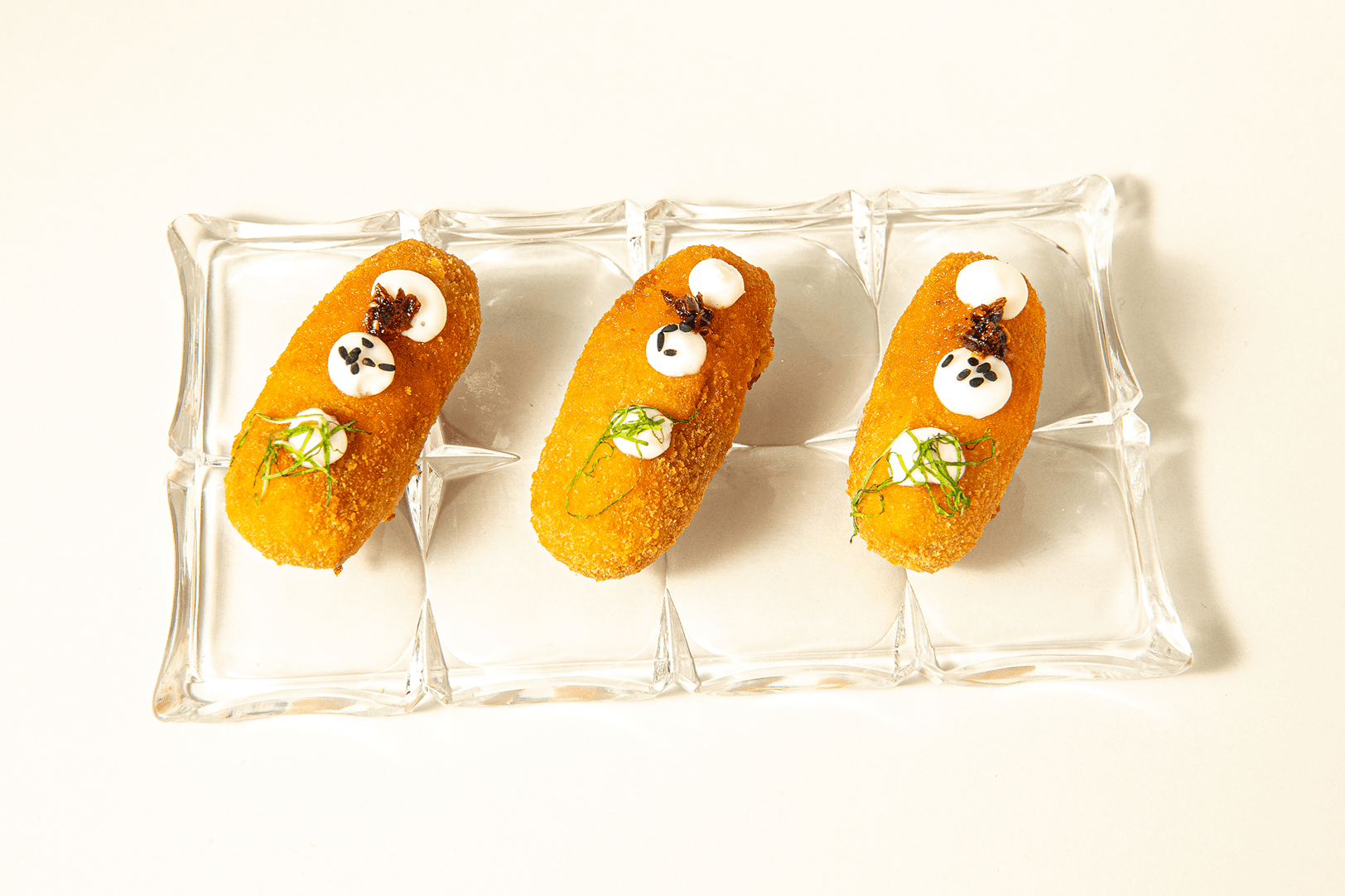 Three golden croquettes lined up on a pressed-glass tray, topped with labneh, black sesame and chilli jam.