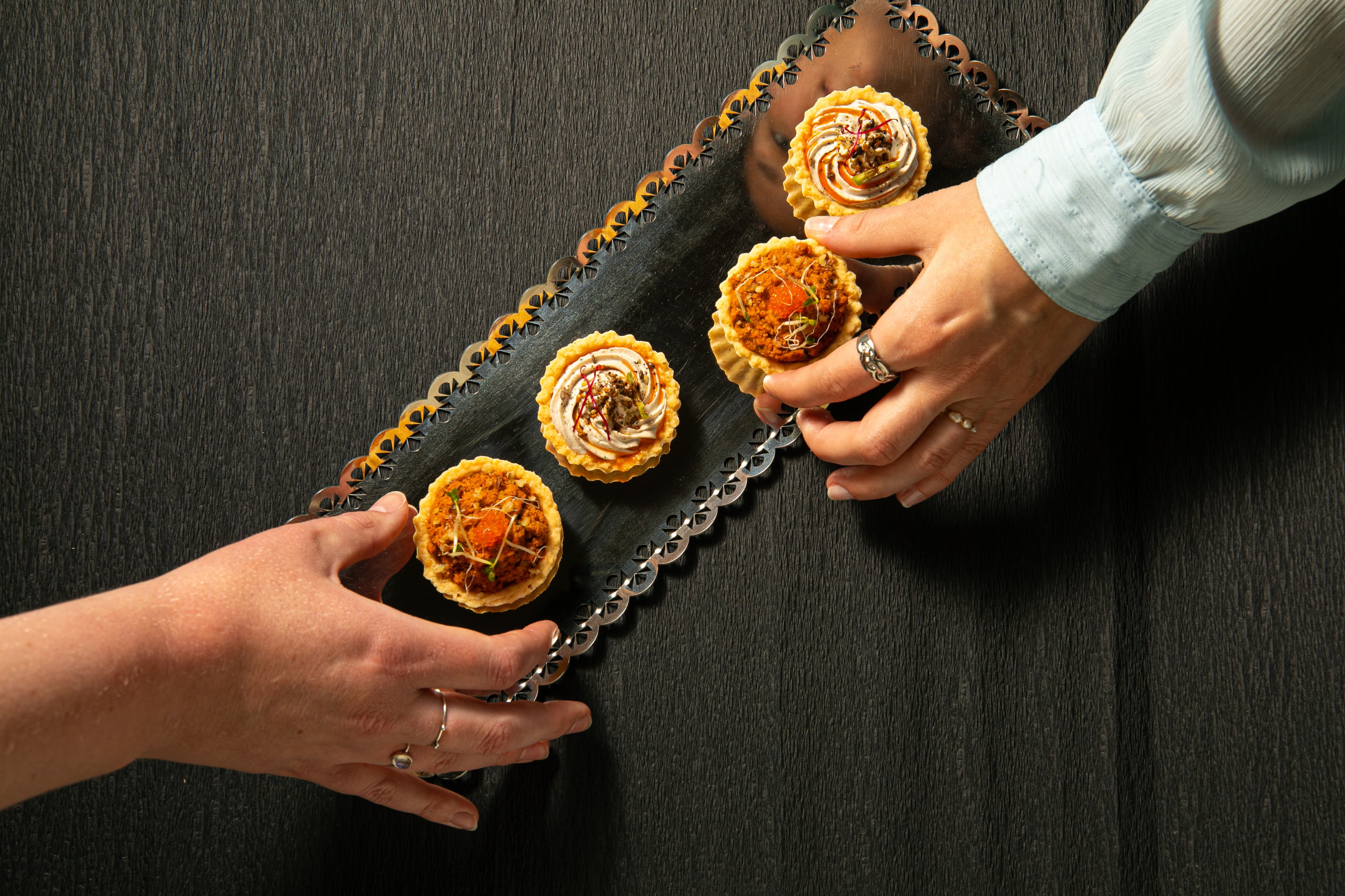 Two pairs of hands lifting savoury tartlets from a scalloped metal tray on a dark wooden table.