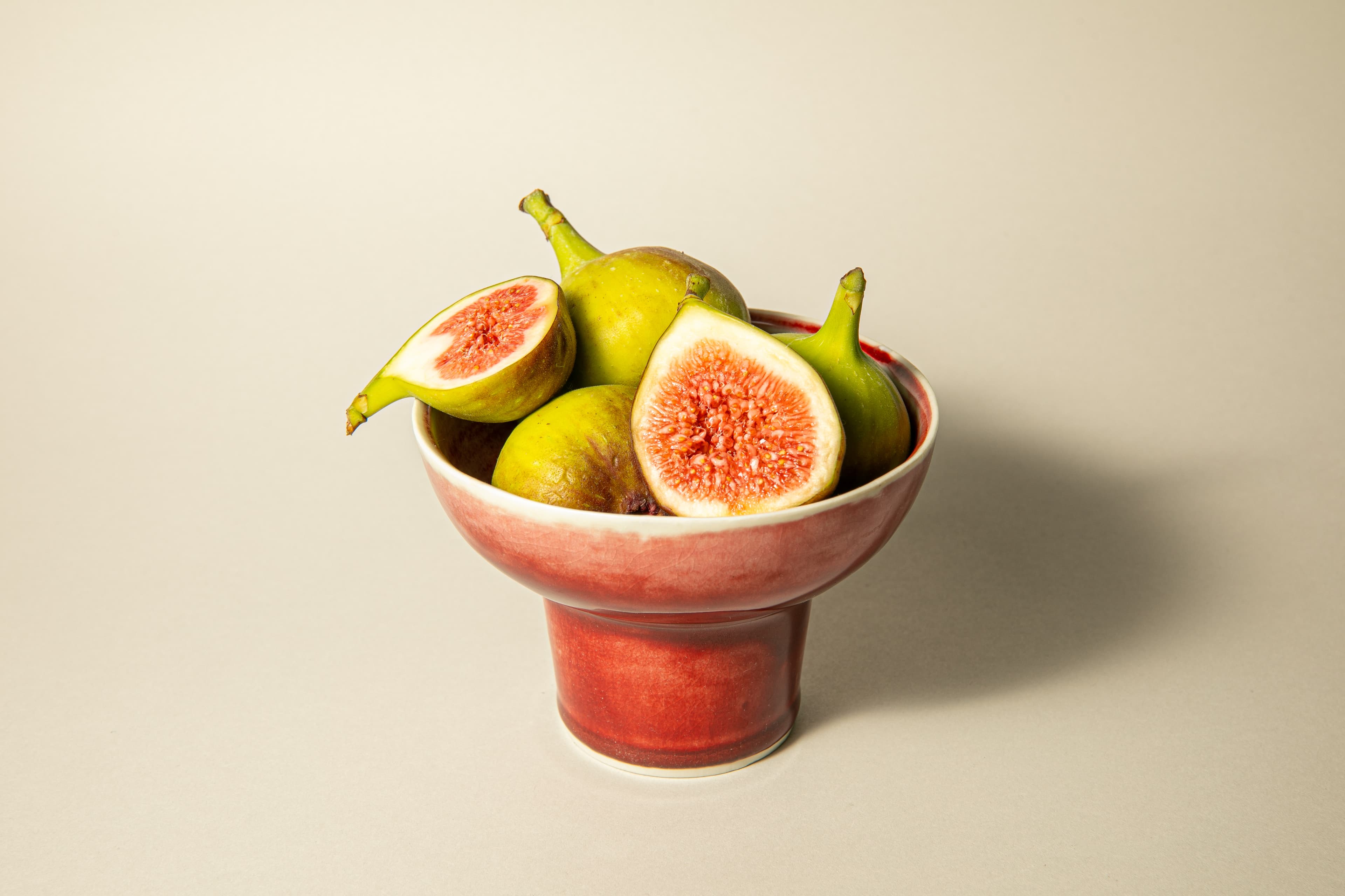 A footed red ceramic bowl holding fresh whole and halved green figs on a soft neutral background.